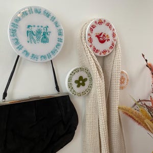 Op de afbeelding: Een verzameling decoratieve wandhaken met vintage bordontwerpen. Eén haak toont een turquoise en wit patroon met figuren, een andere een rood vogelontwerp en een derde een groene bloem. Een zwarte handtas hangt aan een haak en een crèmekleurige sjaal is over een andere gedrapeerd.