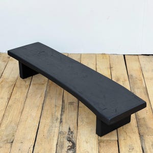 May include: A black wooden bench with a curved seat and two rectangular legs. The bench is painted black and appears to be made of solid wood. The bench is approximately 48 inches long.