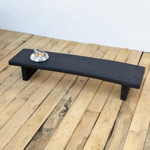 May include: A black wooden bench with a slightly curved top and two supporting legs. A small decorative plate with a seashell is on the bench. The bench is on a wooden floor.