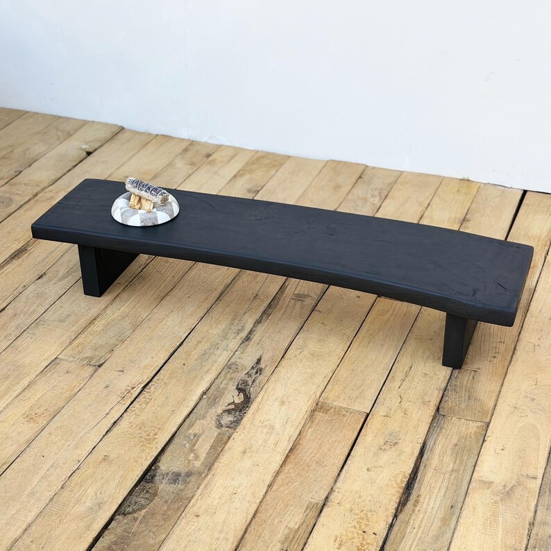 May include: A black wooden bench with a slightly curved top and two supporting legs. A small decorative plate with a seashell is on the bench. The bench is on a wooden floor.