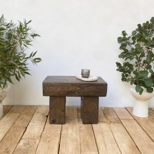 May include: A dark brown, rectangular wooden side table with two square legs. A small, grey glass and a white oval tray sit on top. The table is made of thick, solid wood and has a natural, rustic appearance.
