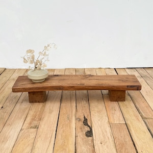 May include: A rustic, rectangular wooden shelf with a natural finish, supported by two square wooden blocks. A small, light-colored ceramic vase with dried floral stems sits on the shelf. The shelf is displayed on a wooden plank floor.