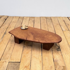Peut inclure: Une table basse en bois faite à la main, de forme naturelle et organique. La table est faite de bois aux tons chauds et repose sur deux supports rectangulaires. Un petit plateau rond en métal avec un objet décoratif est posé sur la table.