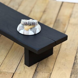 May include: A black wooden altar table with a rectangular top and a single leg. A decorative ceramic dish with a gray and white pattern holds a bundle of sage and a stick of palo santo.