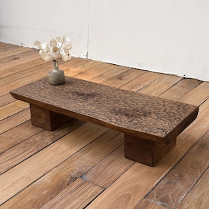 Op de afbeelding: Een lage rechthoekige houten tafel met een donkerbruine afwerking en een getextureerd, kuiltjesoppervlak. De tafel wordt ondersteund door twee dikke blokpoten en bevat een kleine, lichtgekleurde keramische vaas met gedroogde judaspenning.