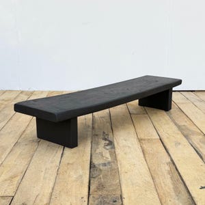 May include: A black wooden bench with a slightly curved seat. The bench is supported by two rectangular legs. The wood has a matte finish. The bench is approximately 60 inches long.