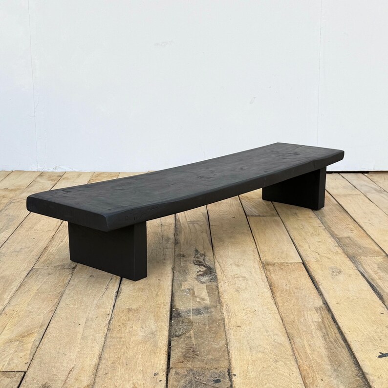 May include: A black wooden bench with a slightly curved seat. The bench is supported by two rectangular legs. The wood has a matte finish. The bench is approximately 60 inches long.