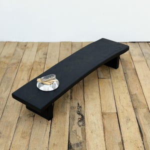 May include: A black wooden bench with a curved top. A small, round, decorative dish with a gray and white pattern holds a bundle of sage and several small wooden sticks. The bench is supported by two legs.