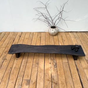 May include: A long, low-profile wooden bench with a dark, charred finish and a natural live edge. The bench features a flat, elongated surface supported by two short, sturdy legs. A small dark teapot and two matching cups sit on the right side.