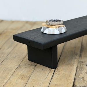 May include: A black wooden bench with a rectangular top and a single leg support. A decorative bowl with a black, white, and gray pattern holds a bundle of sage and several sticks of palo santo.