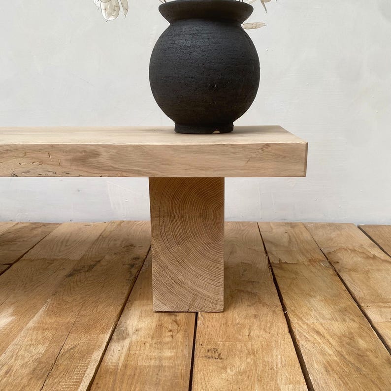 May include: A light-colored wooden bench with a thick, rectangular top and a single, solid leg. A black, textured vase sits on the bench, and dried flowers are visible. The bench is on a wooden floor.