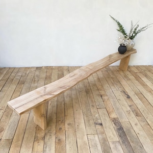 May include: A light-colored wooden bench with a natural, live edge design. The bench is supported by two rectangular legs. A small vase with greenery and a black vase are on the bench.