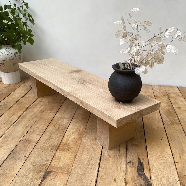 May include: A light-colored wooden bench with a rectangular top and two supporting legs. A small black vase with dried flowers sits on the bench. The bench is made of natural wood and has a rustic appearance.