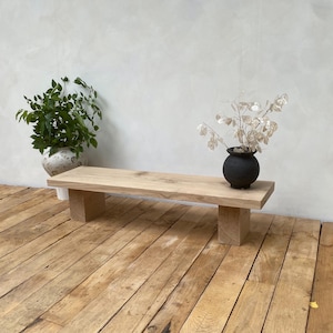 May include: A natural wood bench with a rectangular top and two square supports. A small black vase with dried flowers sits on the bench. A potted plant with green leaves is in the background. The bench is on a wooden floor.