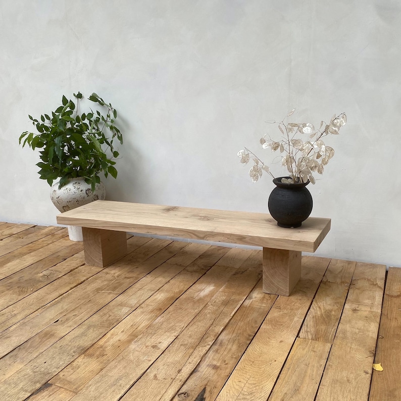 May include: A natural wood bench with a rectangular top and two square supports. A small black vase with dried flowers sits on the bench. A potted plant with green leaves is in the background. The bench is on a wooden floor.