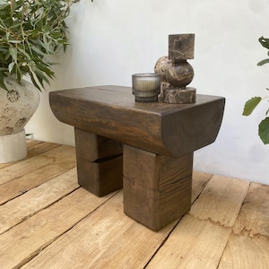 Puede incluir: Mesa auxiliar de madera marrón oscuro con tapa rectangular y dos patas de bloque cuadrado. Sostiene una vela en un portavelas de vidrio y una escultura de mármol. La mesa es de madera maciza. Mide unos 60 cm de largo.