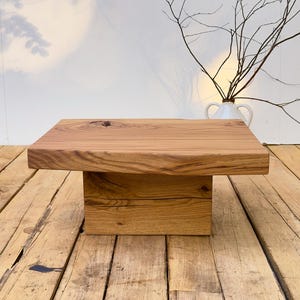 Peut inclure: Une table basse rectangulaire en bois brun clair naturel avec un grain apparent et un nœud visible sur le plateau. La table se compose d'un plateau épais et massif reposant sur un pied central robuste en forme de bloc rectangulaire.