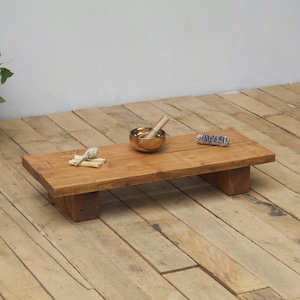 Könnte beinhalten: Ein rechteckiger Meditationstisch aus Holz mit natürlichem Finish. Eine kleine Messing-Klangschale mit einem Holzschlägel, ein Salbeibündel und ein kleiner, gerollter Gegenstand befinden sich auf dem Tisch. Der Tisch wird von zwei kurzen, rechteckigen Stützen getragen.