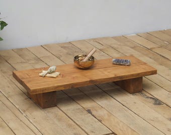 May include: A rectangular, wooden meditation table with a natural finish. A small brass singing bowl with a wooden striker, a bundle of sage, and a small, rolled item are on the table. The table is elevated on two short, rectangular supports.