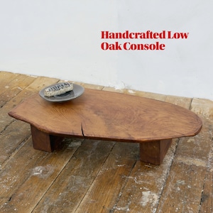 Pode incluir: Consola baixa de carvalho artesanal com acabamento natural de grão de madeira. A mesa tem uma forma orgânica e assenta em dois suportes retangulares. Um prato pequeno com um objeto repousa sobre a mesa. O texto "Handcrafted Low Oak Console" é vermelho.