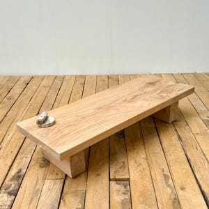 May include: A low rectangular table crafted from light-coloured natural wood with a visible grain pattern and knots. The table features a thick slab top supported by two sturdy block-style legs. A small circular stone object rests on the surface.