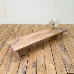 May include: A low rectangular bench crafted from natural light brown wood with a live edge top and two block-style legs. A small, round, light-colored vase containing dried, metallic-toned foliage sits on the right side of the bench surface.