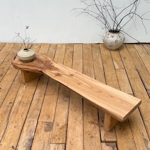 Peut inclure: Un banc long et bas fabriqué à partir d'une seule pièce de bois naturel clair avec un bord brut, soutenu par deux pieds en bois robustes. Un petit vase en céramique ronde de couleur claire contenant des brindilles sombres sans feuilles.