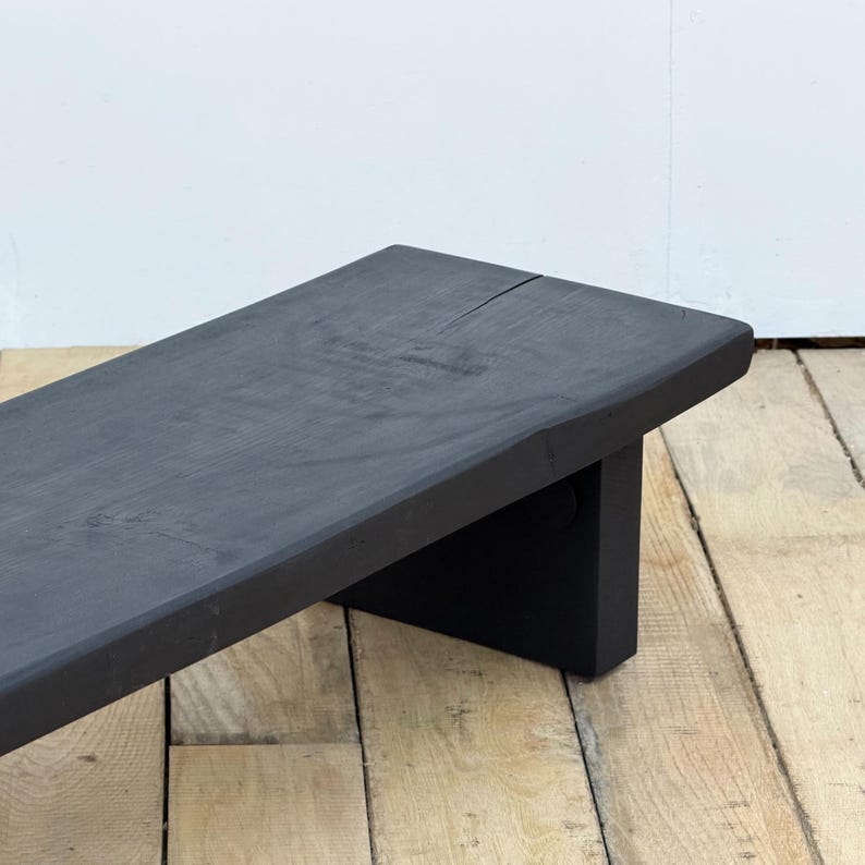 May include: A black wooden bench with a rectangular top and a single leg support. The bench is painted black and has a natural wood grain. The bench is approximately 48 inches long.
