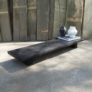 Peut inclure: Une table basse en bois noir avec un plateau rectangulaire et deux pieds courts. La table présente un motif de grain de bois naturel. Deux vases et un livre sont posés sur la table.