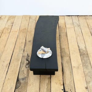 May include: A black, rectangular wooden bench with a curved edge. A small, round, decorative plate with a gray and white pattern holds a bundle of sage and two sticks. The bench sits on a light brown wooden floor.