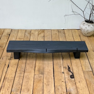 May include: A low, rectangular wooden bench finished in a matte black stain. The bench features a thick, solid top with a visible wood grain texture and two vertical slits along the front edge, supported by two sturdy, matching black wooden legs.