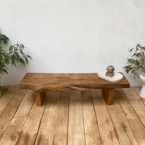 May include: A natural wood bench with a live edge design. The bench is supported by two rectangular legs. A small, decorative stone and a white tray are on the bench. The bench is on a wooden floor.