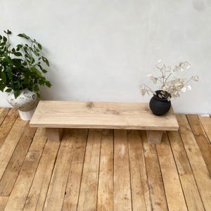 May include: A light-colored wooden bench with a rectangular top and two supporting legs. A small black vase with dried flowers sits on the bench. A white and beige patterned vase with green plants is to the left.