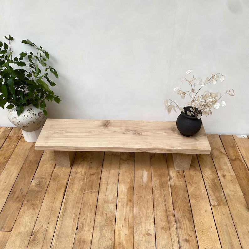 May include: A light-colored wooden bench with a rectangular top and two supporting legs. A small black vase with dried flowers sits on the bench. A white and beige patterned vase with green plants is to the left.