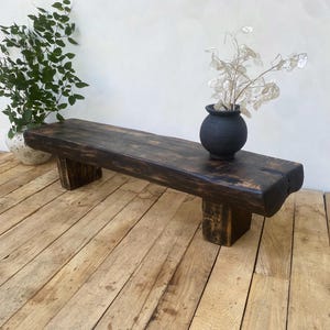 May include: A rustic, dark-stained wooden bench with a natural edge. The bench is supported by two rectangular legs. A small, black vase with dried flowers sits on top. The bench is displayed on a wooden floor.