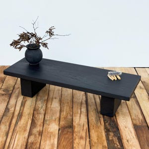 Op de afbeelding: Een zwarte rechthoekige salontafel met een donkere afwerking, ondersteund door twee rechthoekige poten. Een kleine zwarte vaas met gedroogde takken staat op de tafel. Een klein decoratief item rust op het oppervlak van de tafel.