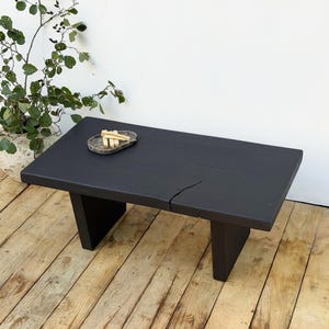 Op de afbeelding: Een zwarte rechthoekige salontafel met een natuurlijke houtnerf. De tafel heeft een scheur over de bovenkant en staat op twee schuine poten. Een kleine, ovale stenen schaal met houten stokjes rust op het tafeloppervlak.