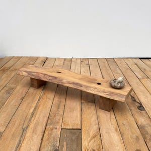 Op de afbeelding: Een handgemaakte houten serveerplank met een natuurlijke rand, ondersteund door twee rechthoekige blokken. Een klein, getextureerd, rond object rust op de plank. Het hout heeft een warme, lichtbruine kleur, op een houten vloer.