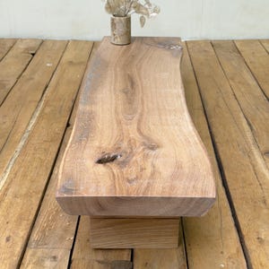 May include: A low wooden bench featuring a natural live edge slab top with visible wood grain and a knot, supported by a rectangular block base. A small cylindrical vase containing dried silver dollar eucalyptus stems sits on the surface.