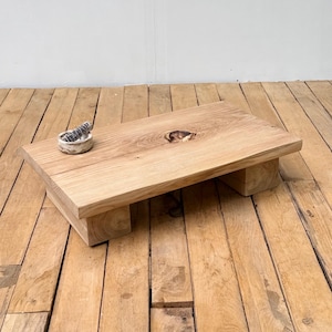 Op de afbeelding: Een lage rechthoekige houten salontafel met een natuurlijke lichtbruine nerf en een opvallende knoest in het midden. De tafel wordt ondersteund door twee dikke blokpoten en heeft een kleine, ronde, lichtgekleurde stenen kom aan de linkerkant.