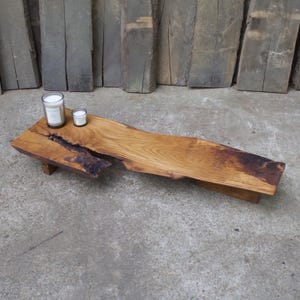 Può includere: Un tavolino da caffè in legno con bordi vivi e forma naturale e organica. Il tavolo è realizzato in legno marrone chiaro con una venatura marrone scuro al centro. Due candele sono appoggiate sul tavolo. Il tavolo è lungo circa 152 cm.