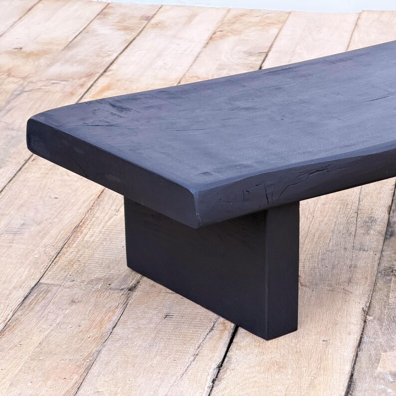 May include: A black wooden bench with a rectangular top and a single, centered support. The bench is painted black and has a slightly rough, textured surface. The wood grain is visible. The bench is set on a light brown wooden floor.