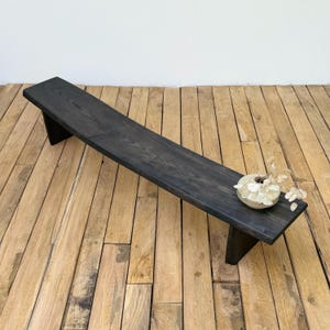 Può includere: Panca in legno tinto scuro dal design minimalista. La panca è rettangolare con un piano piatto e gambe angolate. Un piccolo vaso rotondo color crema con fiori secchi è appoggiato sulla panca. La panca è lunga circa 152 cm.