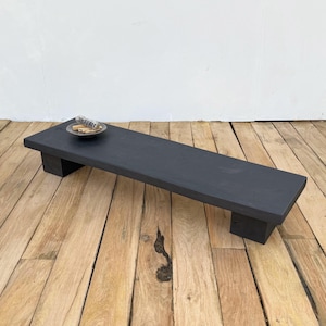 Op de afbeelding: Een lange, rechthoekige, zwarte houten plank met twee korte poten. Een kleine, ronde, zilverkleurige kom met kleine voorwerpen staat op het platform. Het platform staat op een houten vloer.