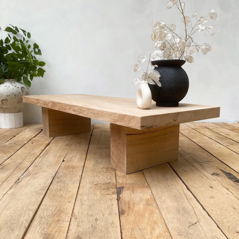 May include: A natural wood coffee table with a rectangular top and two block-style legs. The table is made of light-colored wood and has a minimalist design. A black vase, a white vase, and greenery are on the table.