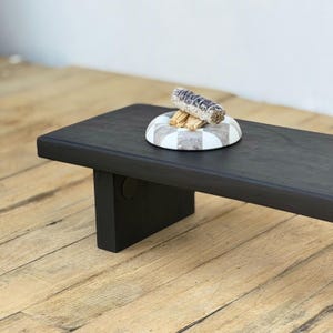 May include: A black wooden altar table with a rectangular top and a single leg. A decorative white and gray checkered dish holds a bundle of sage and some wooden sticks.