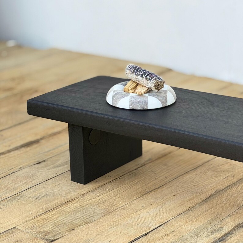 May include: A black wooden altar table with a rectangular top and a single leg. A decorative white and gray checkered dish holds a bundle of sage and some wooden sticks.