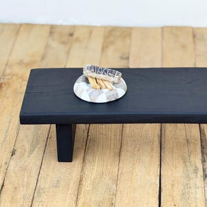 May include: A black wooden bench with a rectangular top and T-shaped legs. A decorative plate with a checkered pattern holds a bundle of sage and several incense sticks. The bench is placed on a wooden floor.