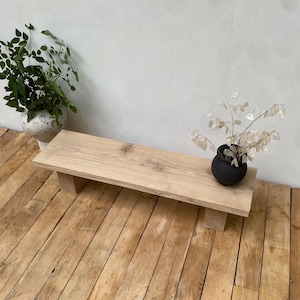May include: A light-colored wooden bench with a rectangular top and four short, square legs. The bench is placed on a wooden floor. Two decorative plants in pots are on the bench and floor.