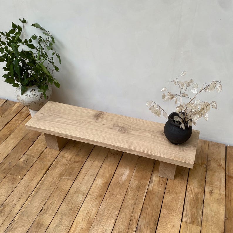 May include: A light-colored wooden bench with a rectangular top and four short, square legs. The bench is placed on a wooden floor. Two decorative plants in pots are on the bench and floor.